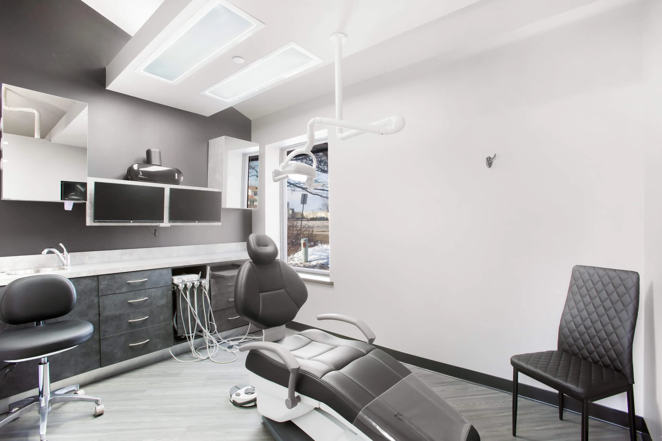Modern dental office with a sleek black dental chair, overhead light, and cabinets on the left. A window offers a view of a snowy landscape.