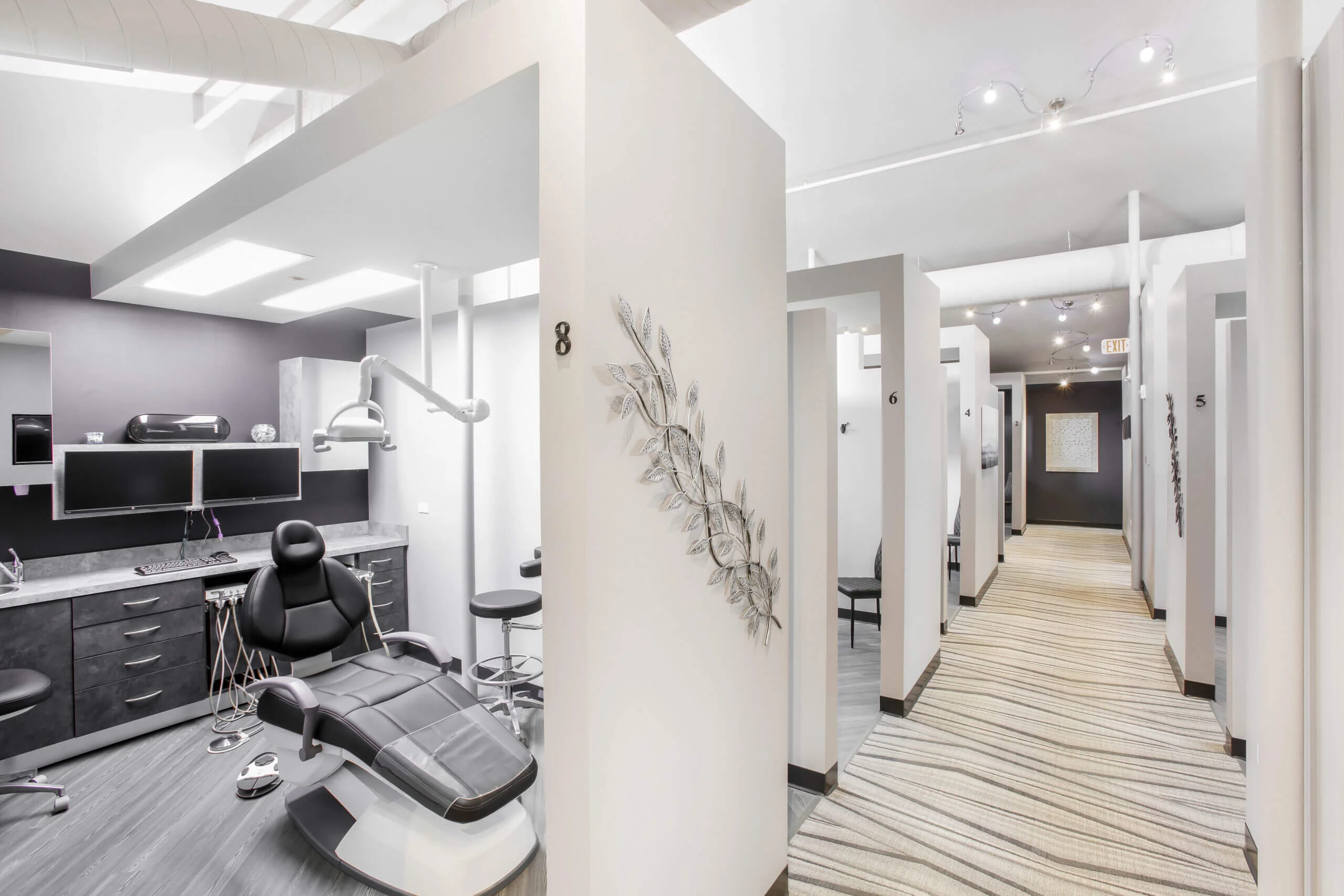 Modern, clean dental clinic hallway with several private treatment rooms. Features sleek black chairs, dual monitors, and bright lighting, conveying a professional and inviting atmosphere.